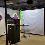 View from the corner of a Revolution One Button Studio. Shown is the AV Media cabinet housing the included tech components. Dual monitors and a camera are mounted on top and a reflective backdrop is hanging near the wall to allow real time background replacement. 